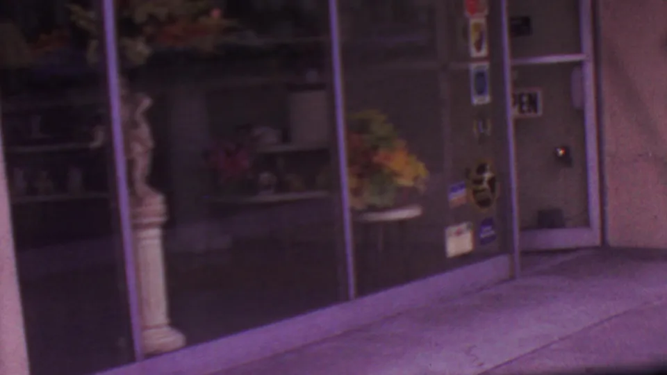 1965 Los Angeles home-movie frame showing reflective glass storefront doors, sidewalk pavement, and a classical-style statue beside the entrance.