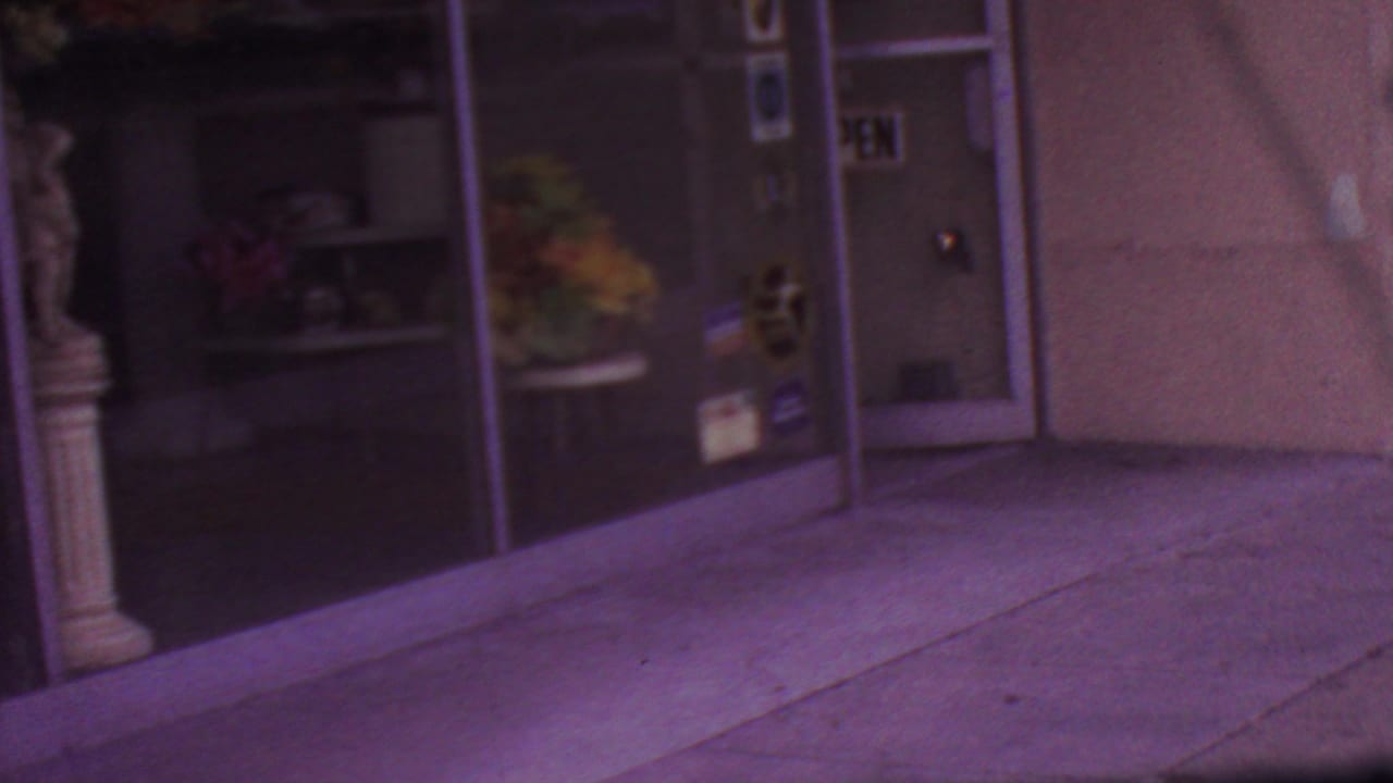 1965 Los Angeles frame showing a glass-fronted storefront entrance with reflective doors, window panels, and sidewalk pavement tinted by the 8mm film color shift.