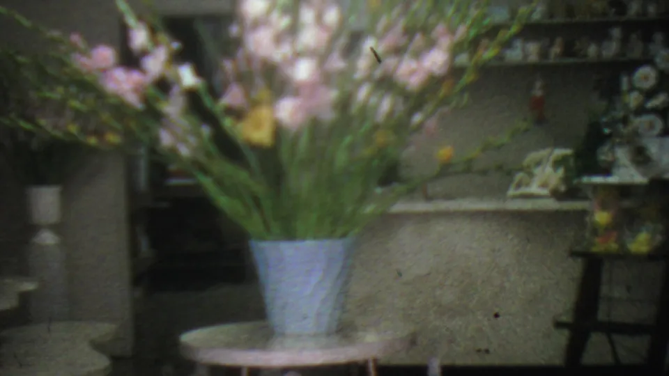 1965 Los Angeles flower shop interior with pink gladiolus and yellow flowers in a pale blue vase, small round tables, and shelves of decorative glassware.