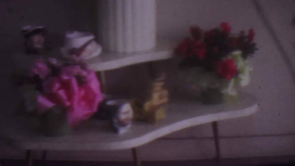 1965 Los Angeles 8mm frame showing a domestic display table with a white planter, pink and red flowers, small figurines, and a softly lit wall.