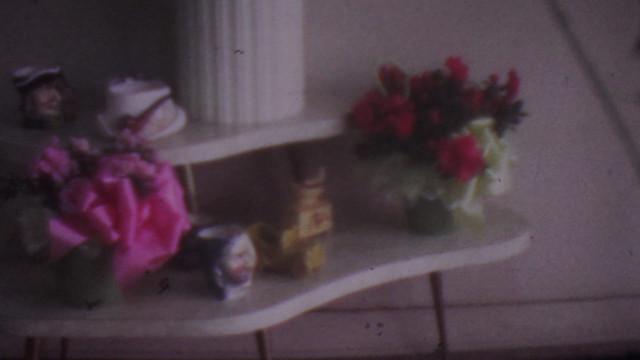 1965 Los Angeles 8mm home-movie frame showing a curved white console table arranged with pink and red flowers, small figurines, white vases, and a fluted column in soft focus.