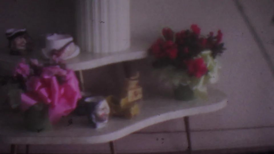 1965 Los Angeles 8mm home-movie frame showing a curved white console table arranged with pink and red flowers, small figurines, white vases, and a fluted column in soft focus.