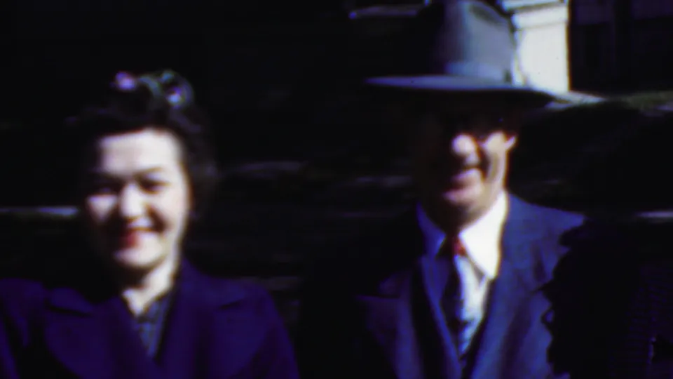In this 1954 Wisconsin 8mm frame, a woman in a dark purple coat smiles beside a man in a suit and broad-brim hat, with another adult partly visible at the edge.