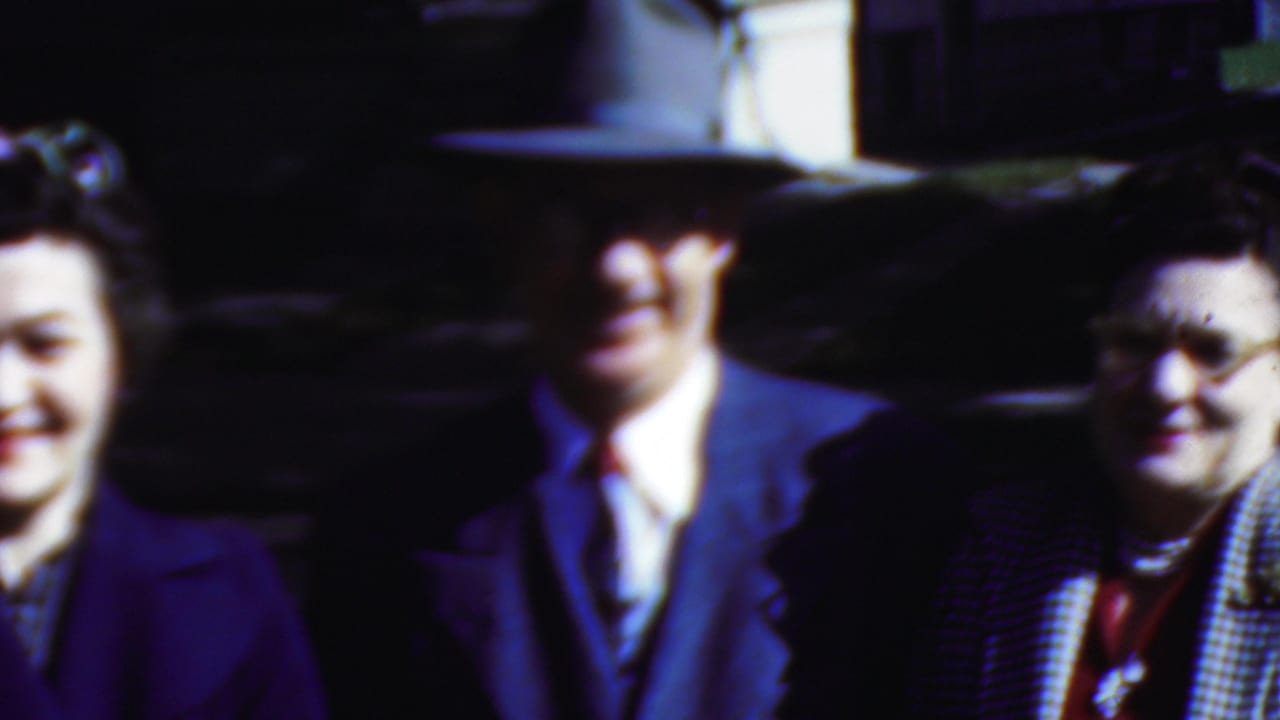Man in a suit and hat stands between two women, one in a purple coat and one in glasses.