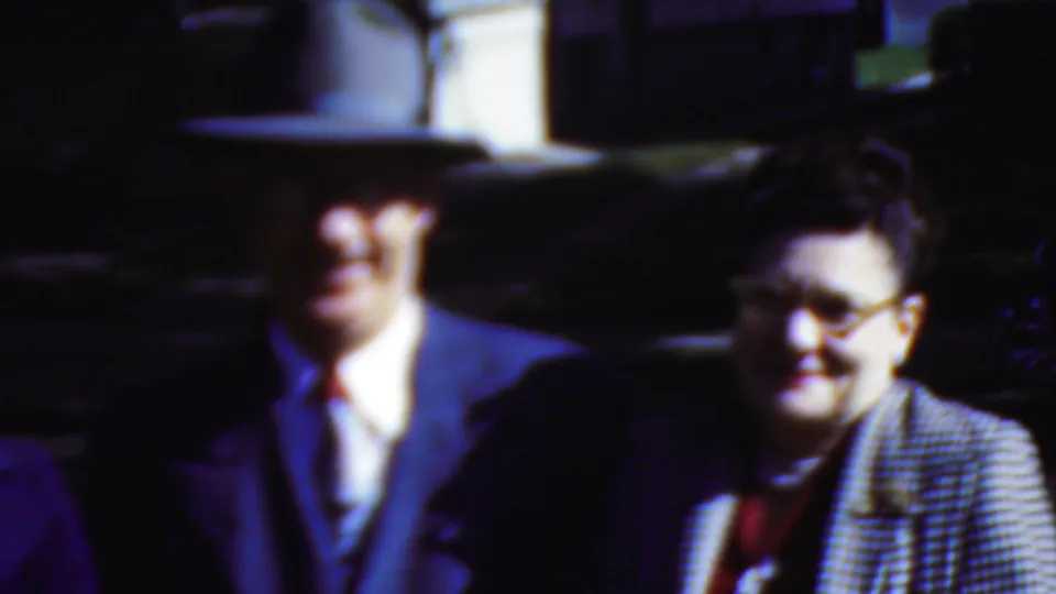 This 1954 Wisconsin 8mm frame shows a man in a suit and hat beside a woman wearing glasses and a checkered jacket, both close to the camera in mixed sunlight and shadow.