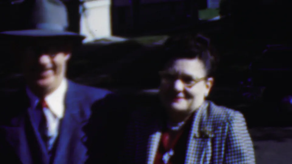In the final 1954 Wisconsin frame, the suited man in a broad-brim hat remains beside the smiling woman in glasses and a checkered jacket against a dark outdoor background.