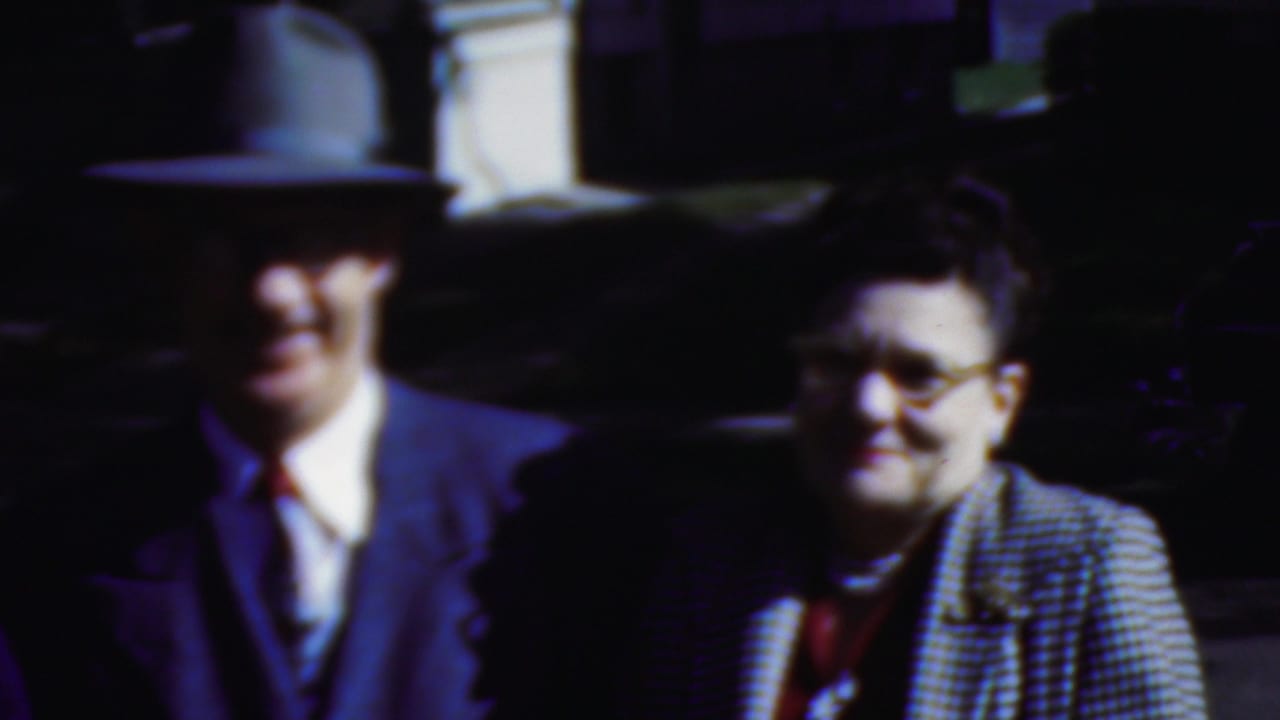 In Wisconsin 1954 home-movie footage, a man in a fedora and suit stands near a smiling woman wearing glasses and a checkered coat as they look toward activity off camera.