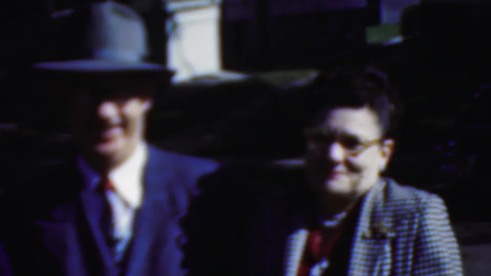 In Wisconsin 1954 home-movie footage, a man in a fedora and suit stands near a smiling woman wearing glasses and a checkered coat as they look toward activity off camera.