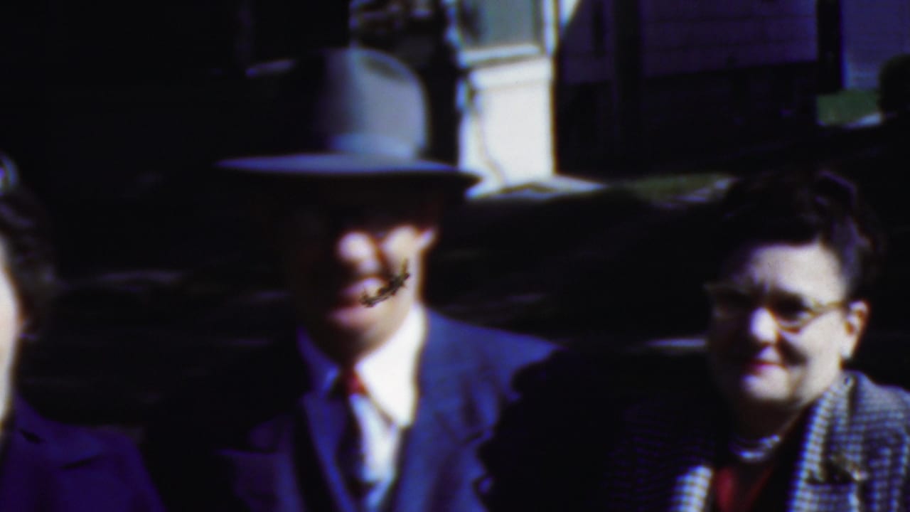 A fedora-wearing man and two adults in formal coats appear in strong sunlight near a dark car, with faces partly softened by motion blur and shadow.