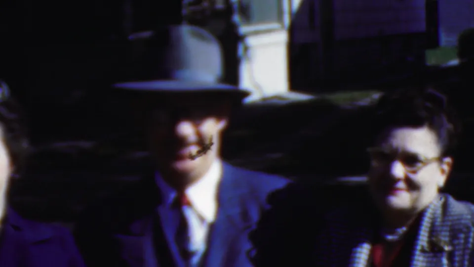 A fedora-wearing man and two adults in formal coats appear in strong sunlight near a dark car, with faces partly softened by motion blur and shadow.