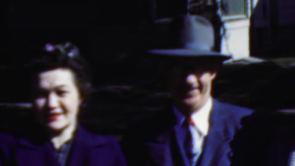 A smiling woman in a dark coat moves closer to the camera while a man in a fedora and suit remains behind her, both framed in a soft 1950s home-movie shot.