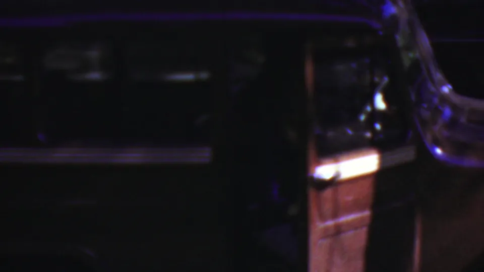 A dark parked automobile occupies most of the frame, with a partially visible figure at the right edge and strong reflections across the vehicle body.