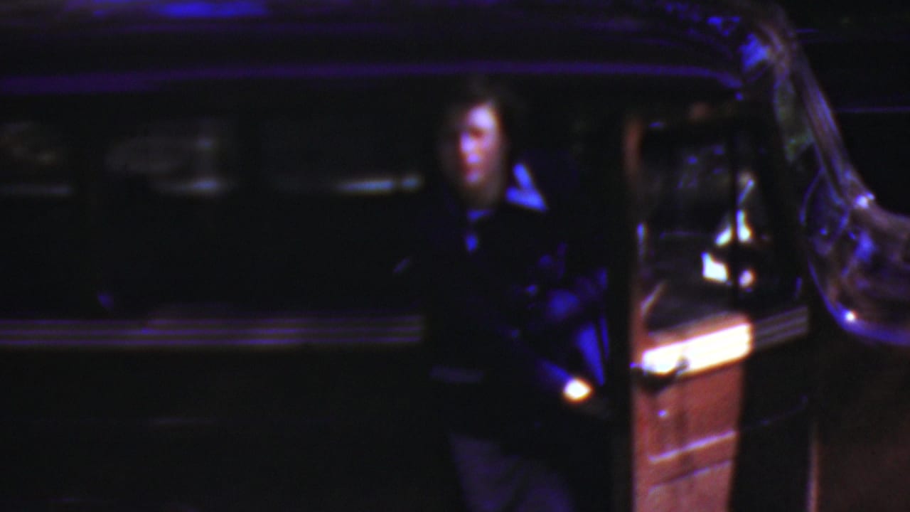 In Wisconsin 1954 home-movie footage, a woman in dark clothing stands near an open vintage car door under very low blue-purple light.