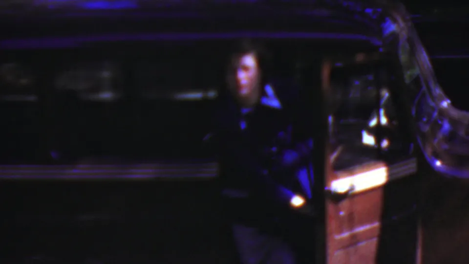 In Wisconsin 1954 home-movie footage, a woman in dark clothing stands near an open vintage car door under very low blue-purple light.