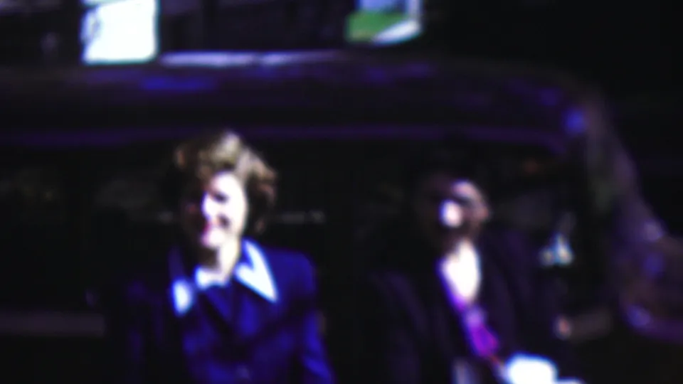 In the supplied 1954 Wisconsin home-movie footage, a woman in a blue suit with a white collar stands beside a dark car at night while other adults are partially visible.