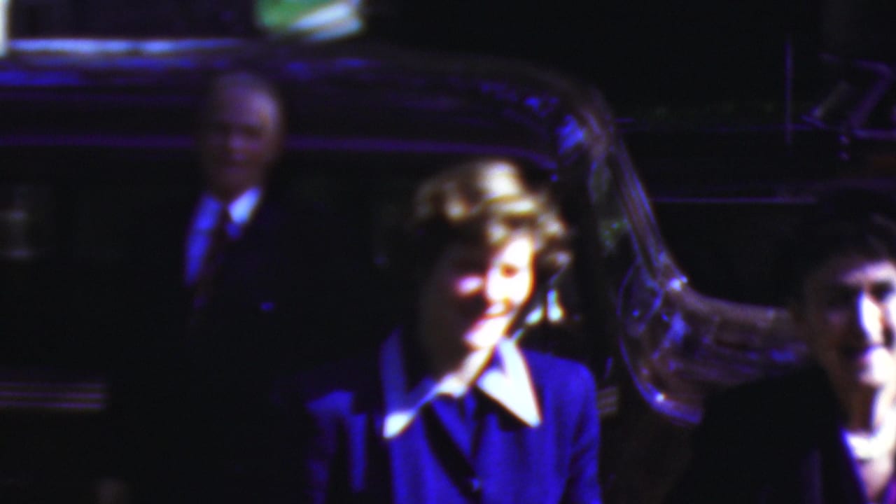 A smiling woman in a blue suit and white collar stands beside a dark car in the nighttime 1954 Wisconsin home-movie scene, with other adults partly visible around her.