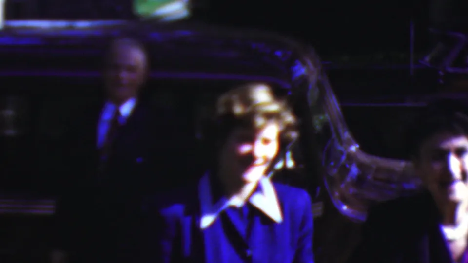A smiling woman in a blue suit and white collar stands beside a dark car in the nighttime 1954 Wisconsin home-movie scene, with other adults partly visible around her.