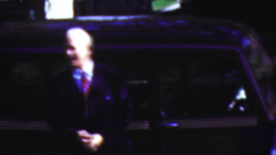 A man in a dark suit and tie stands near a dark classic car in the supplied 1954 Wisconsin home-movie footage, seen in soft nighttime exposure.