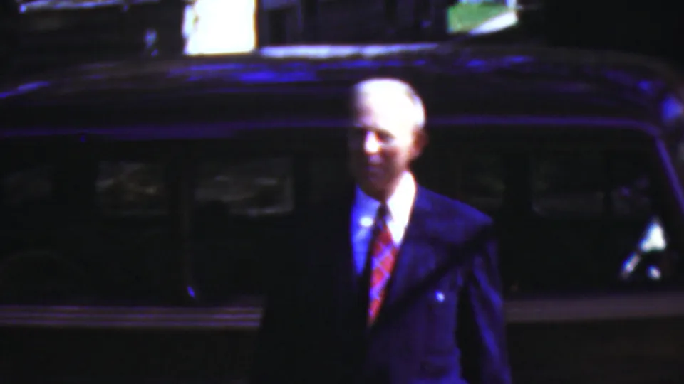 An older man in a dark suit with a red tie stands beside a dark car in the nighttime 1954 Wisconsin home-movie footage, with strong blue-purple color shift across the frame.