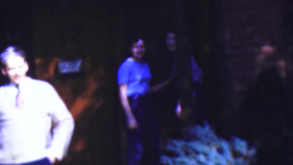 1954 Wisconsin 8mm home-movie frame showing adults in soft focus near a shaded wooded path, with foliage and dark tree cover around them.