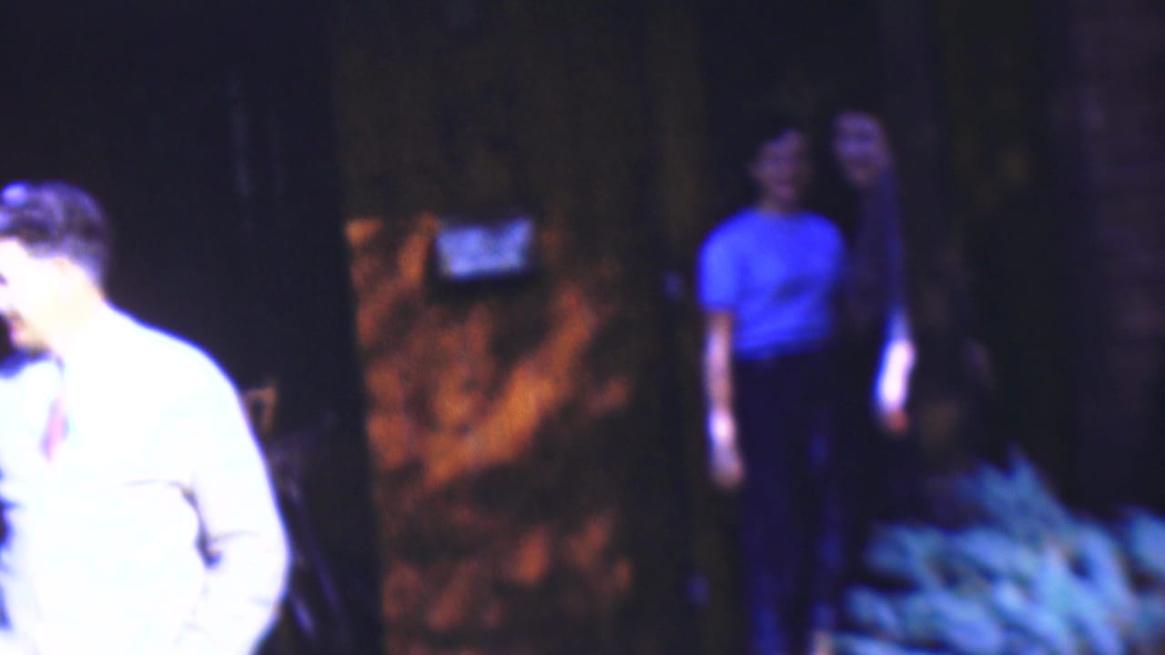 In 1954 Wisconsin home-movie footage, a man in a light shirt and tie is partly visible at the left edge while darker porch or yard details sit behind him.