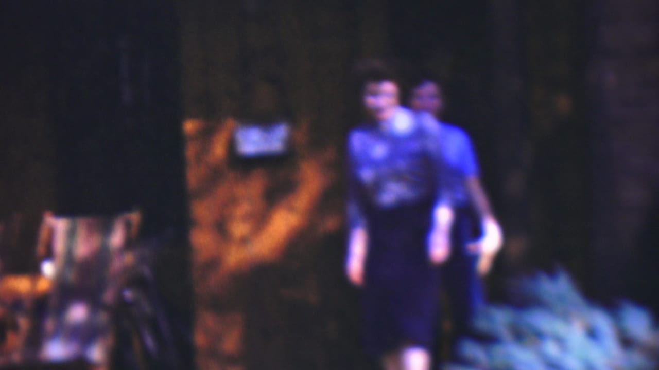 Two adults walk toward the camera near a dark building and foliage.