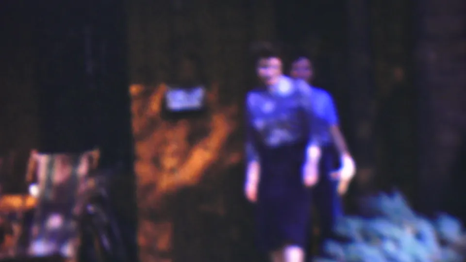 Two adults in blue-toned tops walk toward the camera in a dim outdoor setting with brick or masonry and foliage visible behind them.