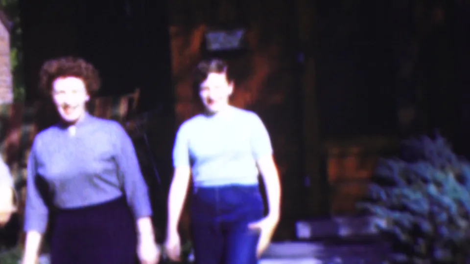 Two women in mid-century dress walk toward the camera in the 1954 Wisconsin home-movie scene, smiling under bright artificial light with foliage and a dark building behind them.