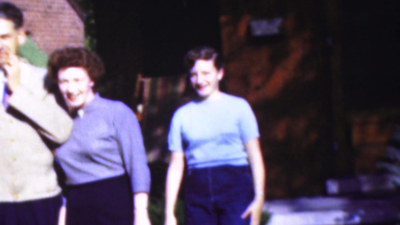 1954 Wisconsin 8mm frame showing three adults outside a brick building, including a woman in a purple blouse, a woman in a light blue blouse, and a partly cropped man.