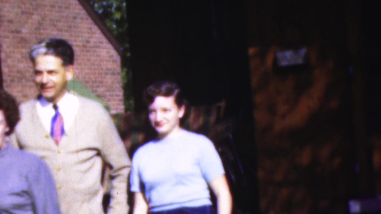 A man in a beige cardigan stands beside a woman in a light blue blouse near a brick building.