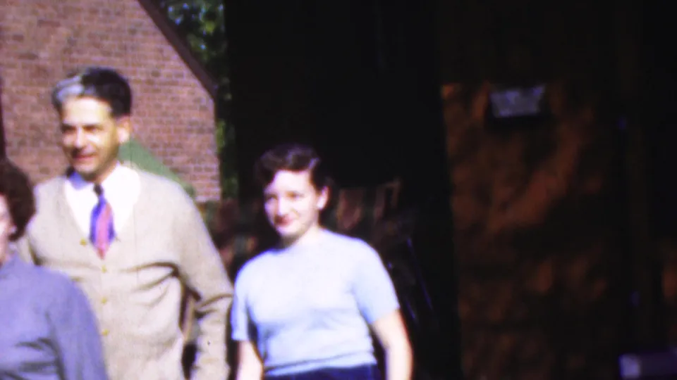 1954 Wisconsin home-movie frame with a smiling man in a beige cardigan and tie standing near a woman in a light blue blouse outside a brick building.