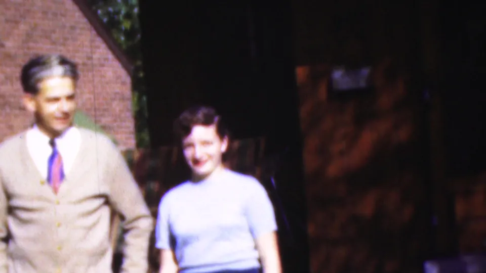1954 Wisconsin 8mm frame showing a man in a beige cardigan and tie beside a smiling woman in a light blue blouse near a shaded brick structure.