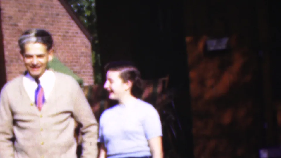 1954 Wisconsin home-movie frame of a man in a beige cardigan and tie smiling beside a woman in a light blue blouse outside a brick building.