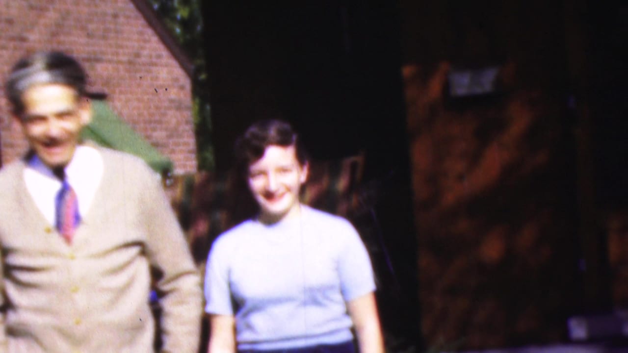 In 1954 Wisconsin home-movie footage, a man in a beige cardigan and tie walks beside a smiling woman in a light blue top near a brick house and green awning.