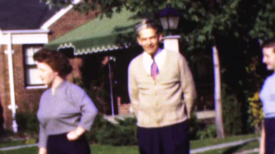A woman in a gray-blue sweater turns across the foreground while a man in a beige cardigan stands near the lawn and green awning outside a brick house in Wisconsin home-movie footage.