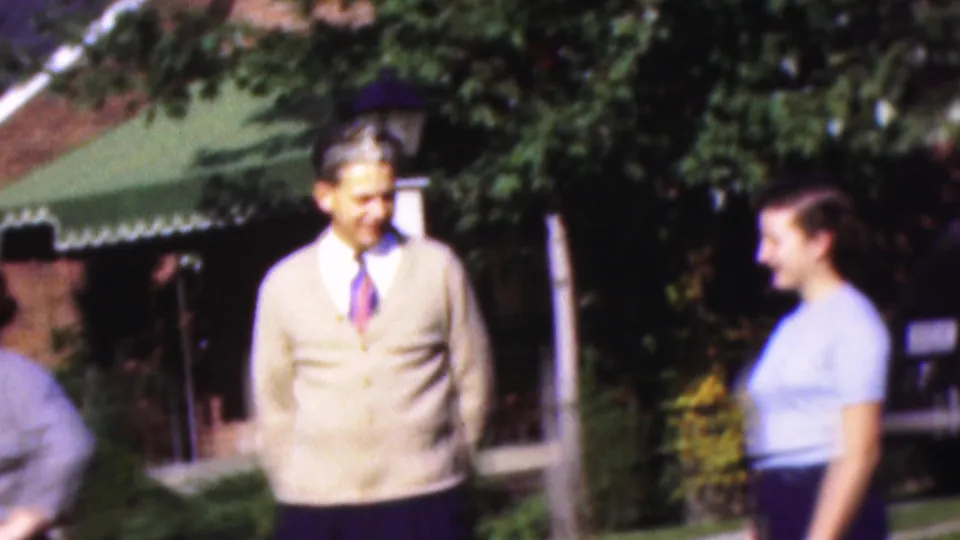 The man in a beige cardigan and the woman in a light blue top face each other near shrubs, a lamp post, and a green awning outside a brick house in 1954 Wisconsin footage.