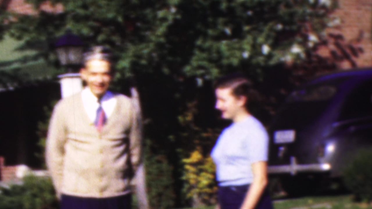 In Wisconsin 1954 home-movie footage, a man in a beige cardigan and tie stands on a lawn while a young woman in a light blue top faces him near a dark parked car and brick house.