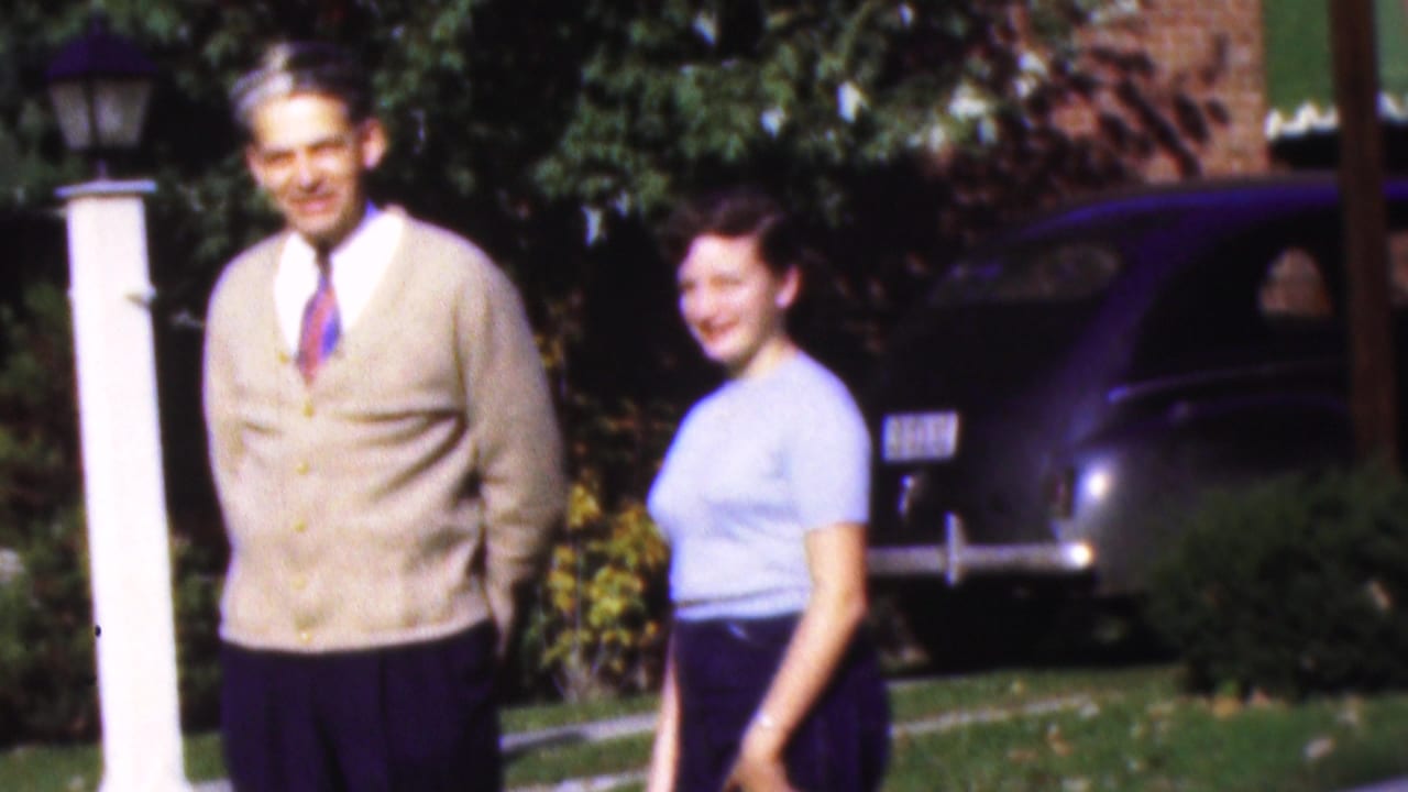 Man and young woman stand on a lawn beside a dark car and house entrance.
