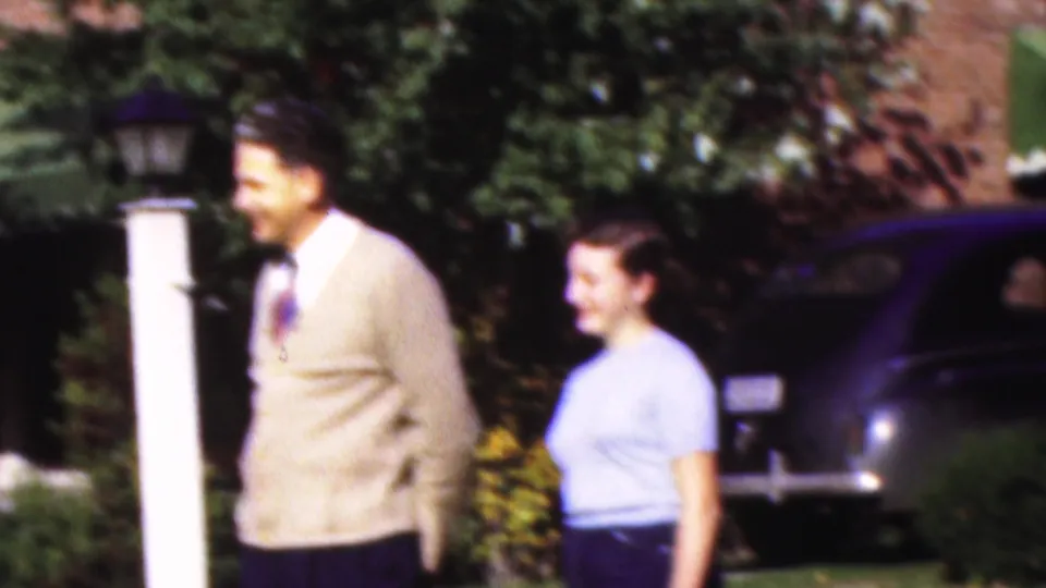 The man turns in profile and the young woman smiles nearby, with a white post lantern, shrubs, dark parked car, and brick house forming the suburban Wisconsin home-movie background.