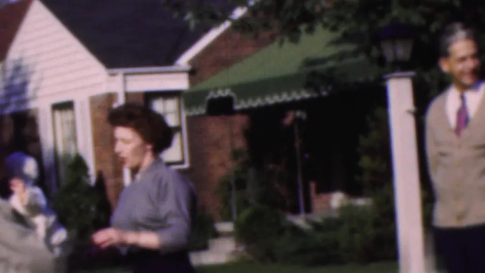 A woman holding a baby appears near the house entrance beneath a green awning, while the man in the beige cardigan is partly visible at the edge of the frame.