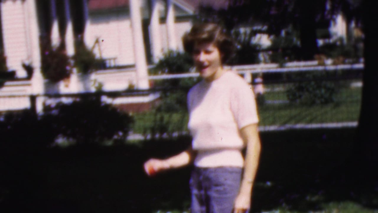 In supplied 1954 Wisconsin home-movie footage, a young woman in a pale pink short-sleeved sweater and blue pants stands on a lawn near a house porch.