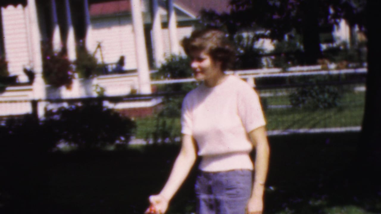Young woman smiling and gesturing in a yard with a porch behind her.