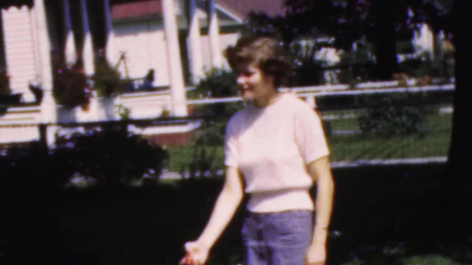 In supplied 1954 Wisconsin footage, the woman smiles while moving one hand in a sunlit yard with porch posts, shrubs, and house siding behind her.