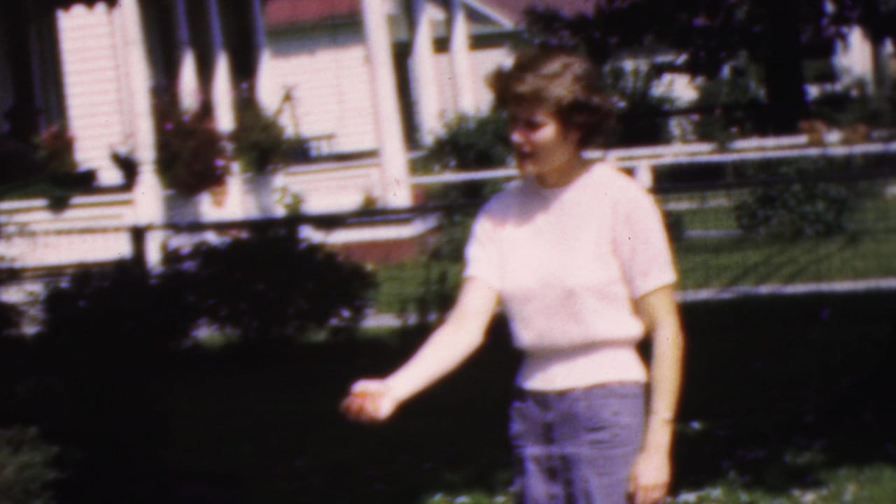 In supplied 1954 Wisconsin home-movie footage, the woman extends a hand as if tossing or offering something while standing in the yard near the house porch.