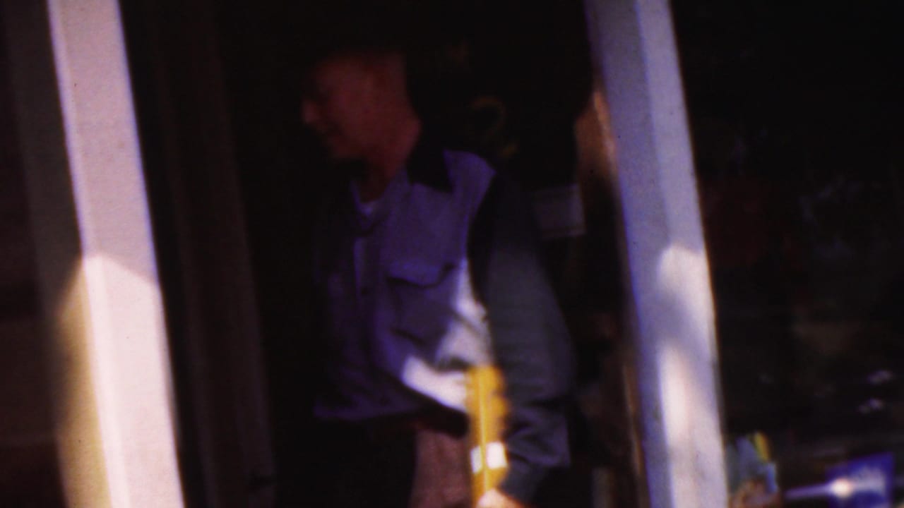 In supplied 1954 Wisconsin footage, a man in a purple and white work-style uniform stands partly in shadow at a doorway while holding a yellow object.