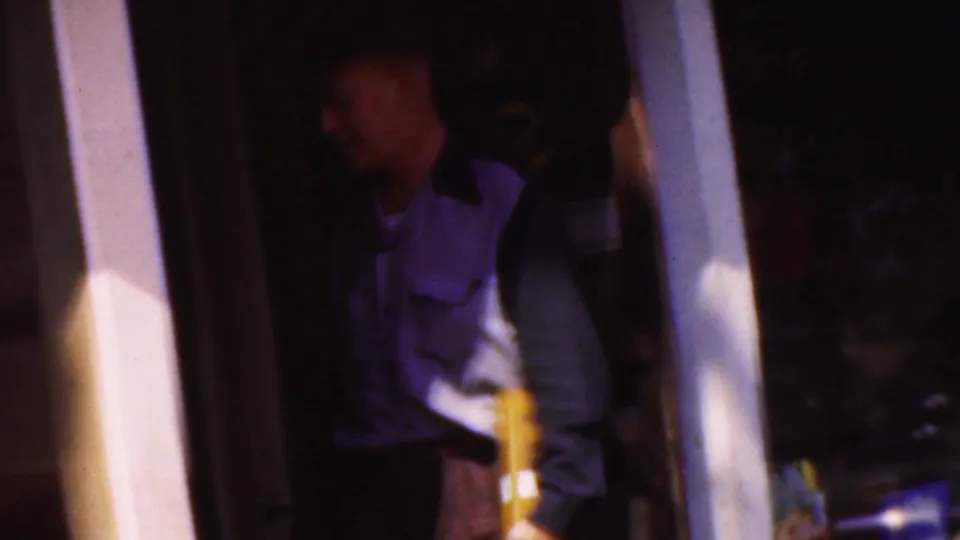 In supplied 1954 Wisconsin footage, a man in a purple and white work-style uniform stands partly in shadow at a doorway while holding a yellow object.