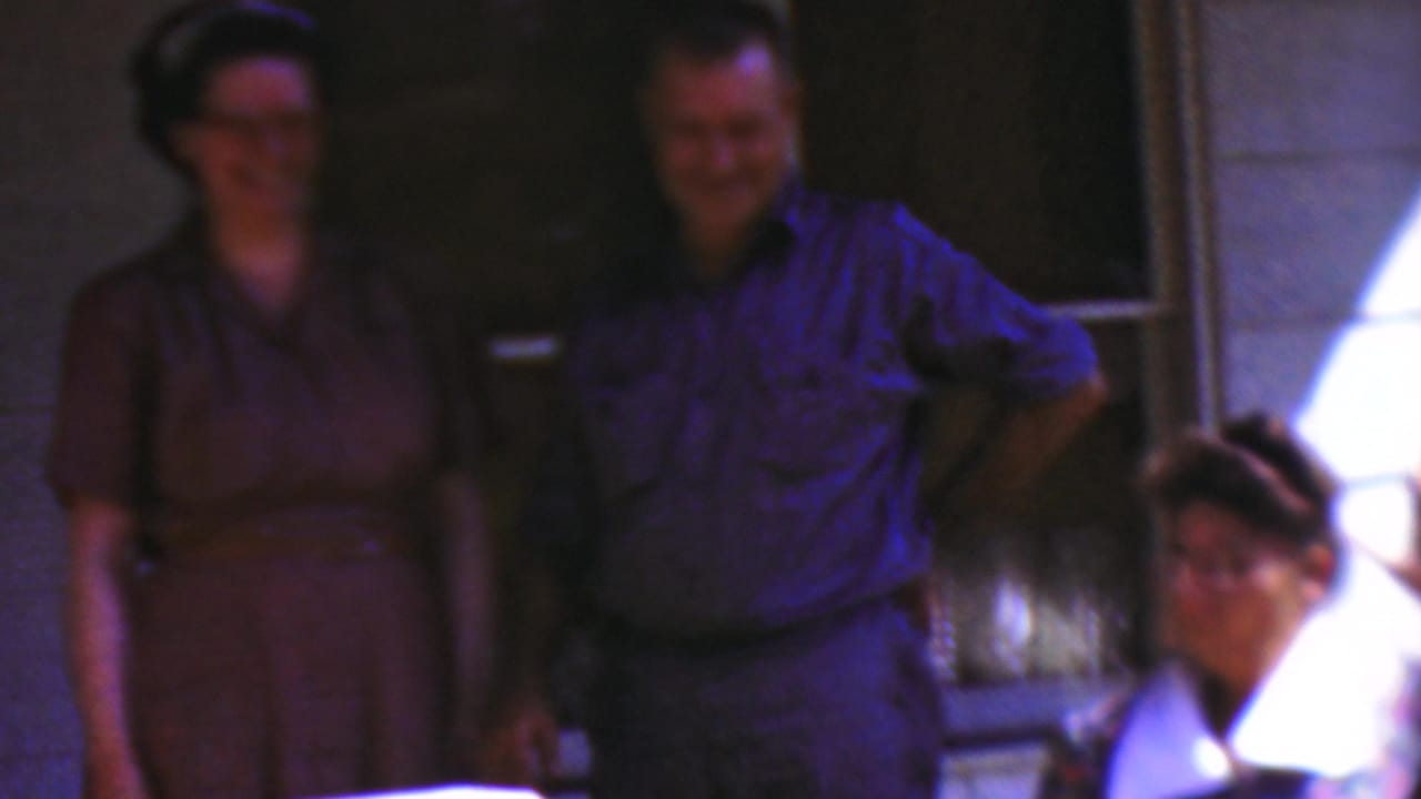 1954 Wisconsin home-movie frame showing a woman in a brown dress and a man in a blue shirt smiling on a porch, with another woman seated in front.