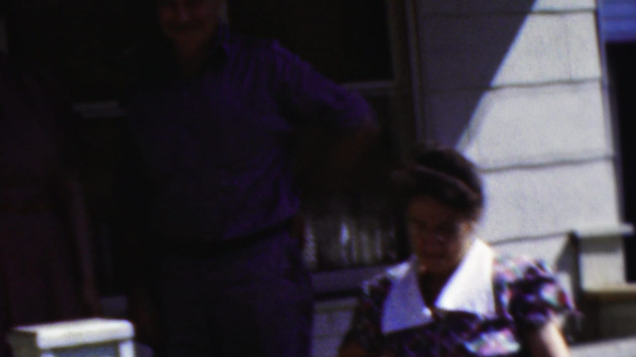 1954 Wisconsin home-movie frame with a seated woman in a patterned dress and white collar near the porch, while standing adults remain partly obscured behind her.