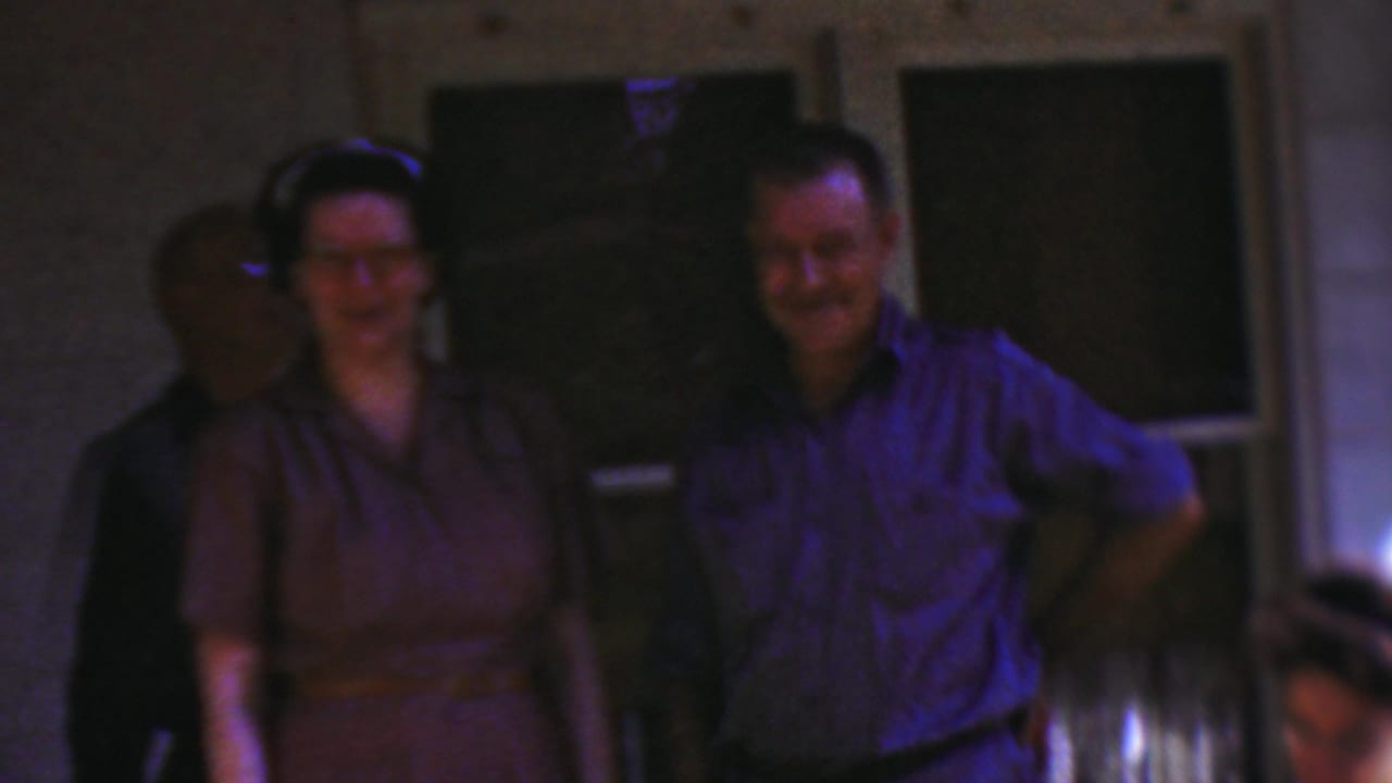 1954 Wisconsin home-movie frame showing a smiling woman in a brown dress and a smiling man in a blue shirt standing together on a residential porch.