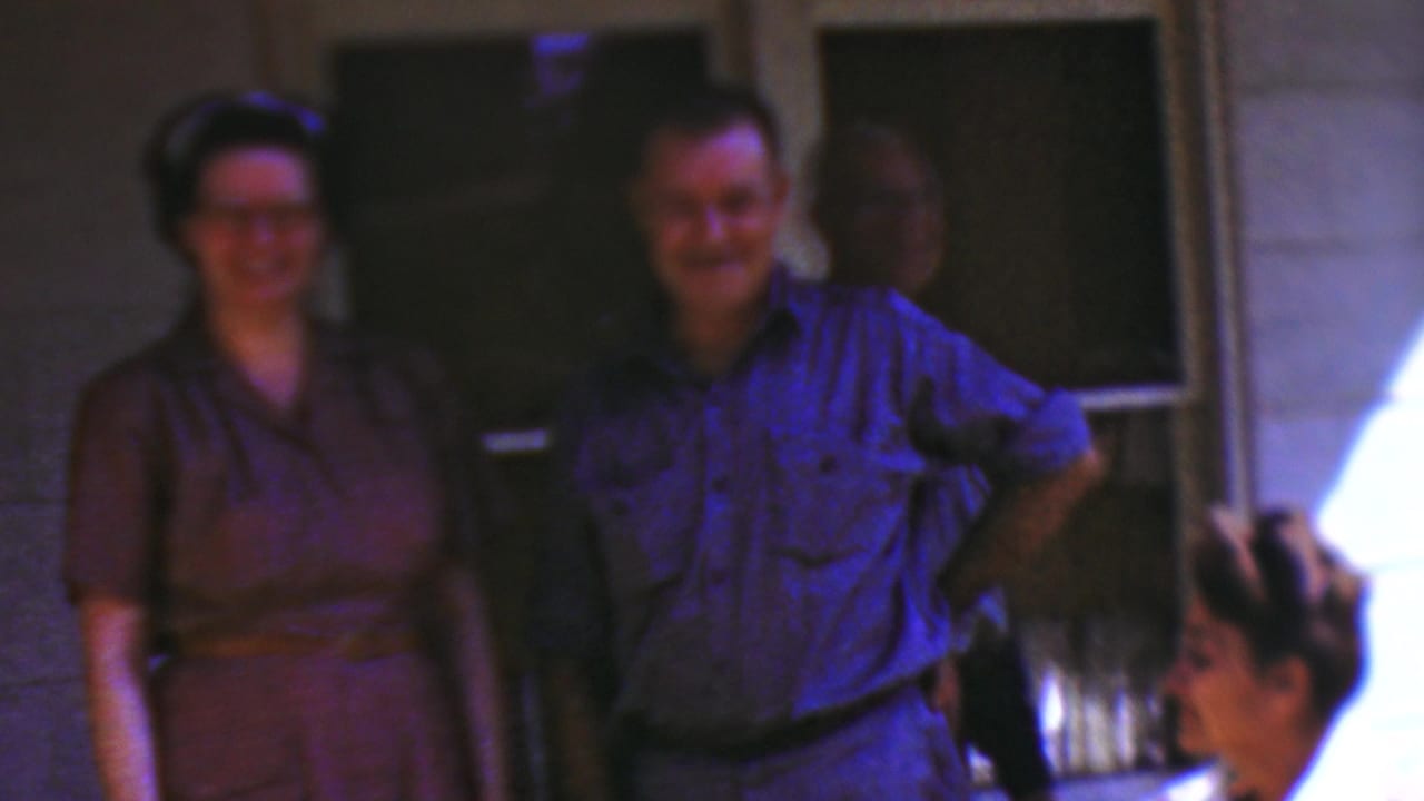 1954 Wisconsin home-movie frame of a woman in a brown dress and a man in a blue shirt smiling on a porch, with another adult standing in shadow and a seated woman partly visible.